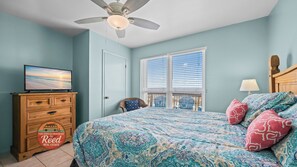 Ein Schlafzimmer mit einem Bett, einem Fernseher auf einer Kommode, einem Deckenventilator und einem Fenster mit Jalousien.