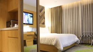 A hotel room with a wooden wardrobe, a bed with white bedding, a television on a wooden cabinet, and a large window with curtains. - HANSA - A Premium Residence (Dhaka)