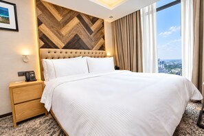 A hotel room with a large bed, a nightstand, a telephone, and a view of the city through the window. - HANSA - A Premium Residence (Dhaka)