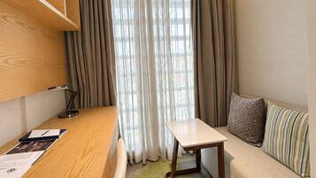 A compact room with a wooden desk, a small table, a sofa, and a window with curtains.