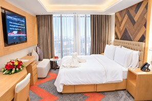 A hotel room with a bed, a desk, a chair, a TV, and a view of the city through large windows. - HANSA - A Premium Residence (Dhaka)