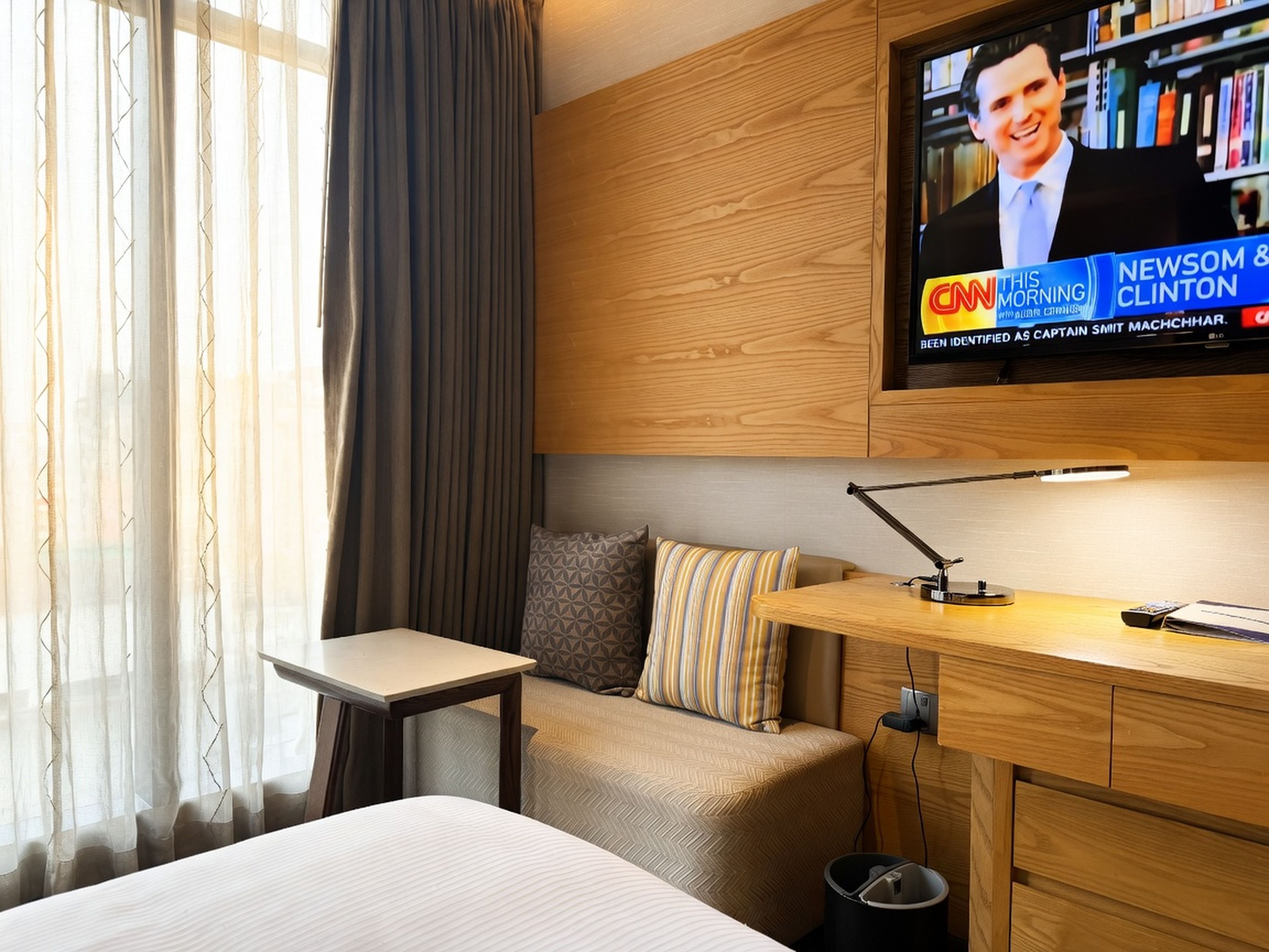 A hotel room with a bed, a desk, a television mounted on the wall, and a window with curtains.