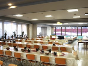 A large conference room with multiple tables and chairs, large windows with curtains, and potted plants along the windowsill.