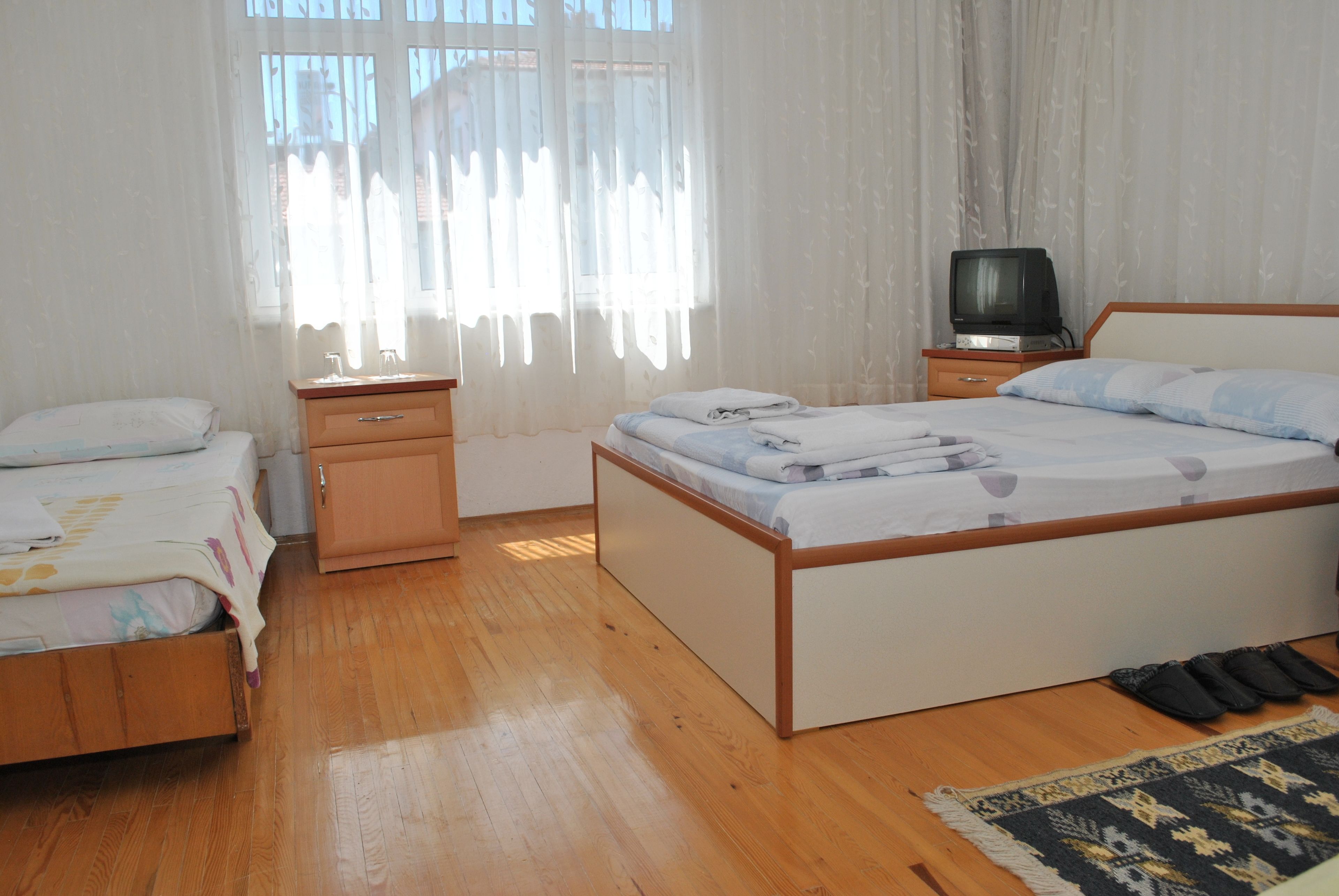 Une chambre avec deux lits simples, un plancher en bois, un téléviseur posé sur une table de chevet et une fenêtre avec des rideaux légers.