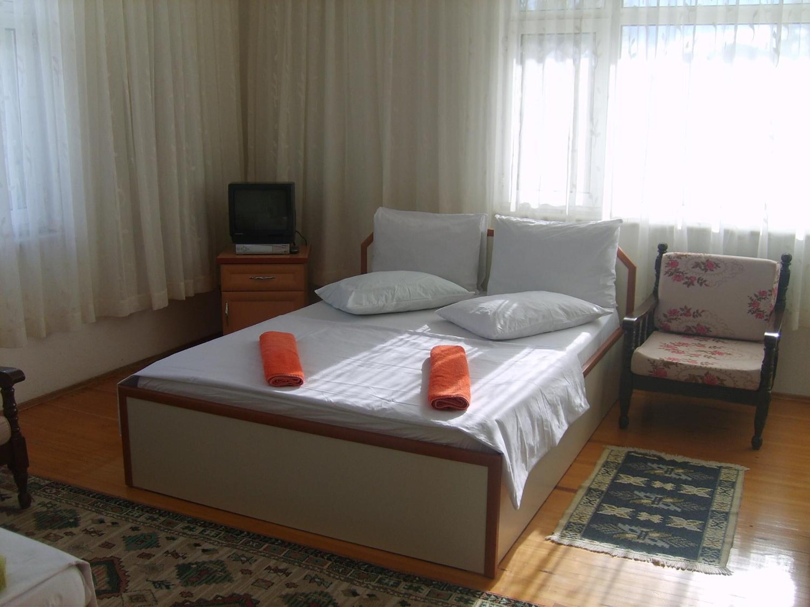 Una cama bien hecha con dos toallas enrolladas, un televisor sobre un soporte de madera, un sillón con estampado floral y una alfombra estampada sobre piso de madera.