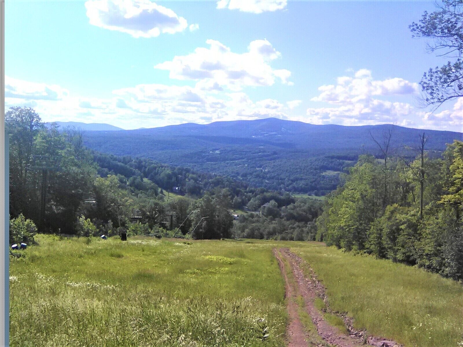 Un champ verdoyant, avec un sentier de terre qui mène vers une vallée bordée d’arbres et dominée par des montagnes lointaines.