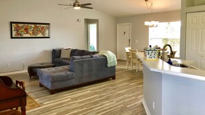 A living room with a gray sectional sofa, a wooden coffee table, and a ceiling fan. There is a dining area with a white table and chairs, and a kitchen with a white countertop and a sink. - Surfside Area Vacation Home Awaits (Cape Coral)