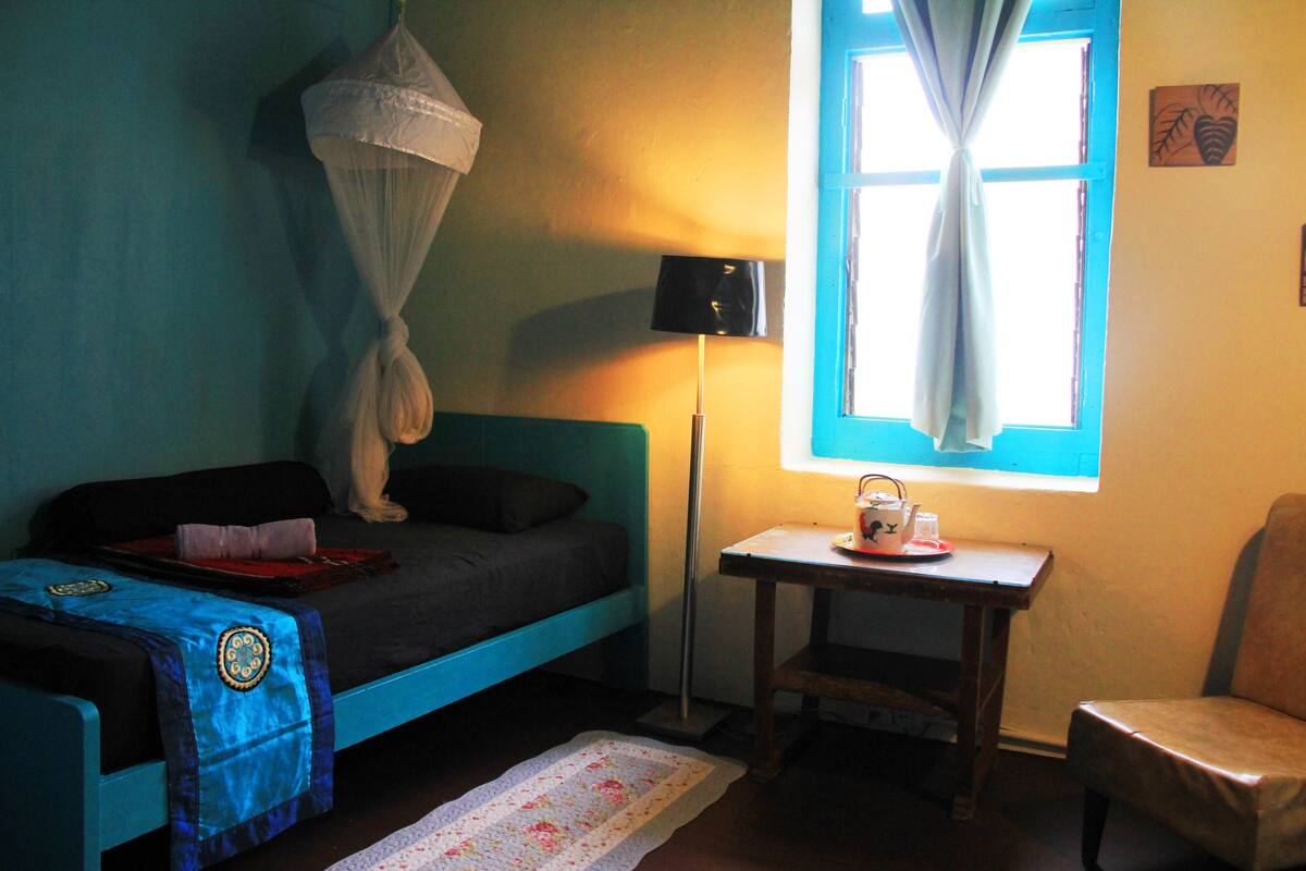 Une pièce comprenant un lit, une petite table, une chaise, une lampe et une fenêtre avec des rideaux.
