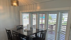 A dining area with a dark wooden table, four chairs, and a candle centerpiece. Large windows with blinds offer a view of outdoor furniture and greenery. - Beach house on private resort (Port Isabel)