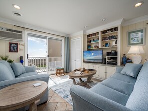 Un salon avec un canapé, une table basse et un téléviseur. Il y a un balcon avec vue, une bibliothèque et un tableau accroché au mur.