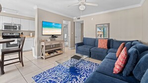 A modern living room with a blue sofa, a glass coffee table, and a kitchen area with white cabinets and a microwave. - 2BR Family Beach Escape – Silver Sands #253S by RVA (Longboat Key)