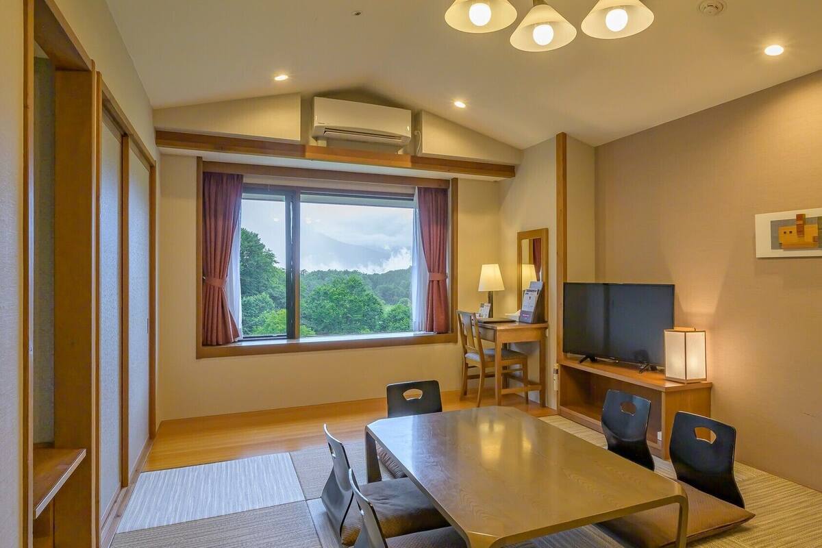 A hotel room with a dining table, chairs, a TV, a desk with a lamp, and a large window with curtains.