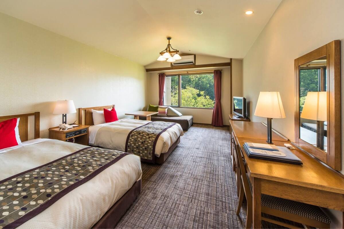 A hotel room with two beds, a desk, a TV, and a window with a view of trees.