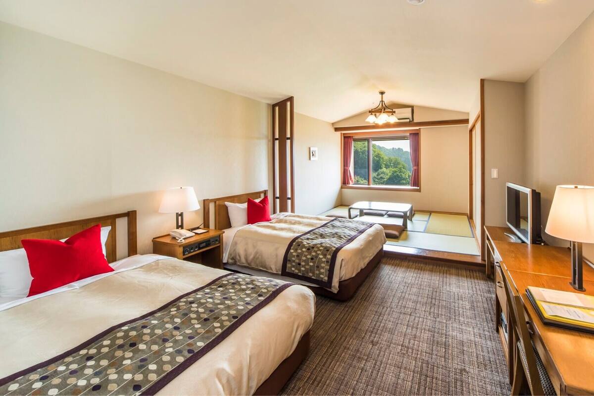 A hotel room with two beds, a desk with a TV, a window with a view, and a ceiling light.