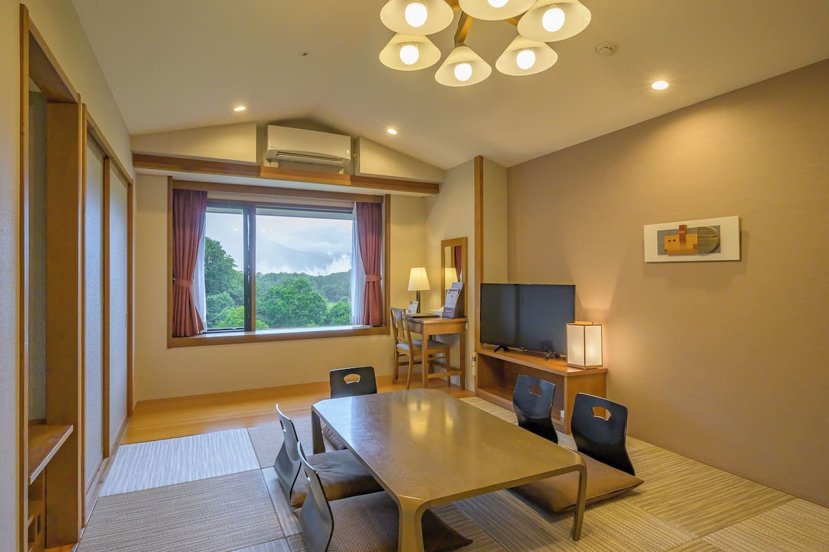 A hotel room with a large window, a dining table, chairs, a TV, and a desk with a lamp.