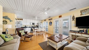 A spacious living room with a wooden floor, a dining area with white chairs, a kitchen with white cabinets, and a large TV on the wall. - Cute beach house w/ramp for easy access. 3Bd/2Ba- Great Gulf Views/easy access. (Gulf Shores)