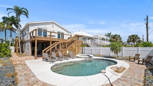 A pool with a wooden deck and lounge chairs, a two-story house with a covered patio, and palm trees in the background.