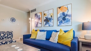 Una sala de estar con un sofá azul, cojines blancos y amarillos estampados, una mesa de centro y cuadros abstractos en la pared.