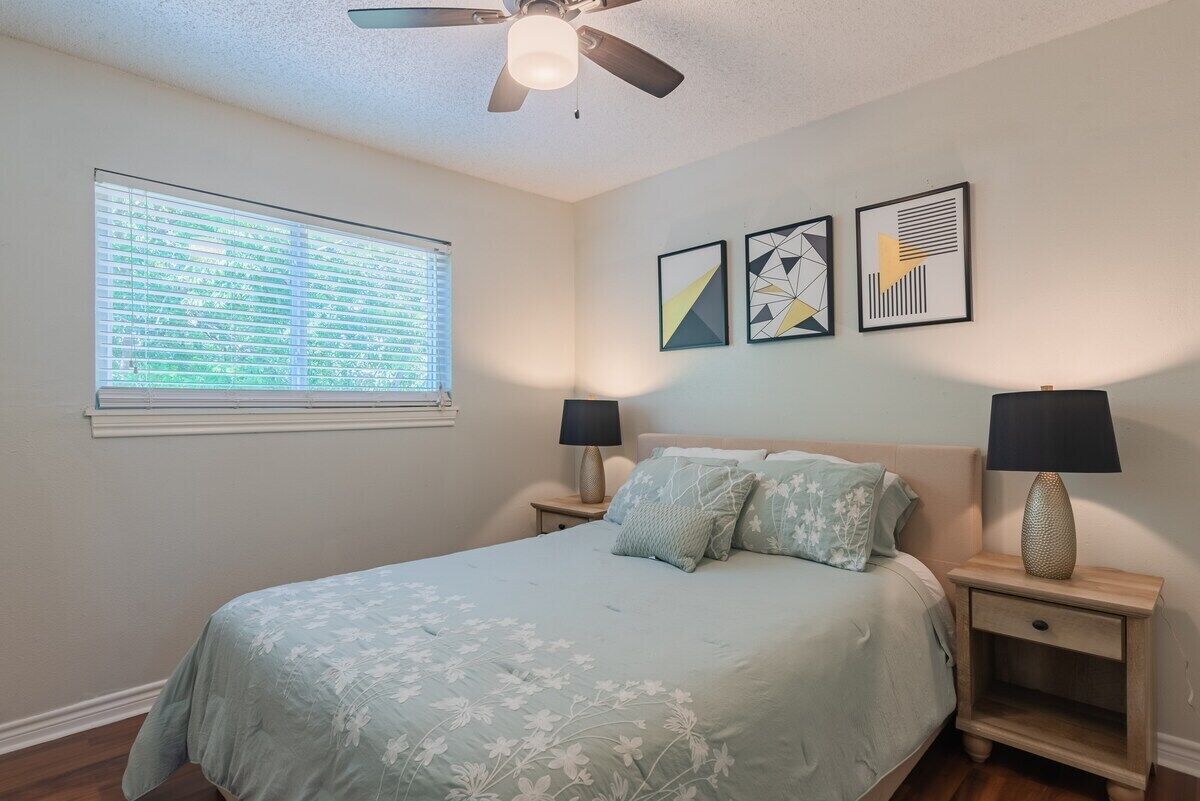 A bedroom with a bed, two bedside tables with lamps, a ceiling fan, and three framed artworks on the wall.