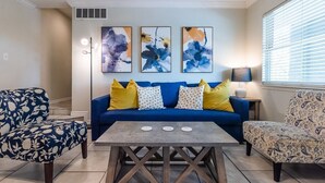 Una sala de estar con un sofá azul, sillas con estampado, una mesa de centro y arte abstracto en la pared.