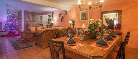 Un comedor con una mesa de madera dispuesta para seis personas, rodeada de sillas. Se ve un árbol de Navidad con luces de colores al fondo.