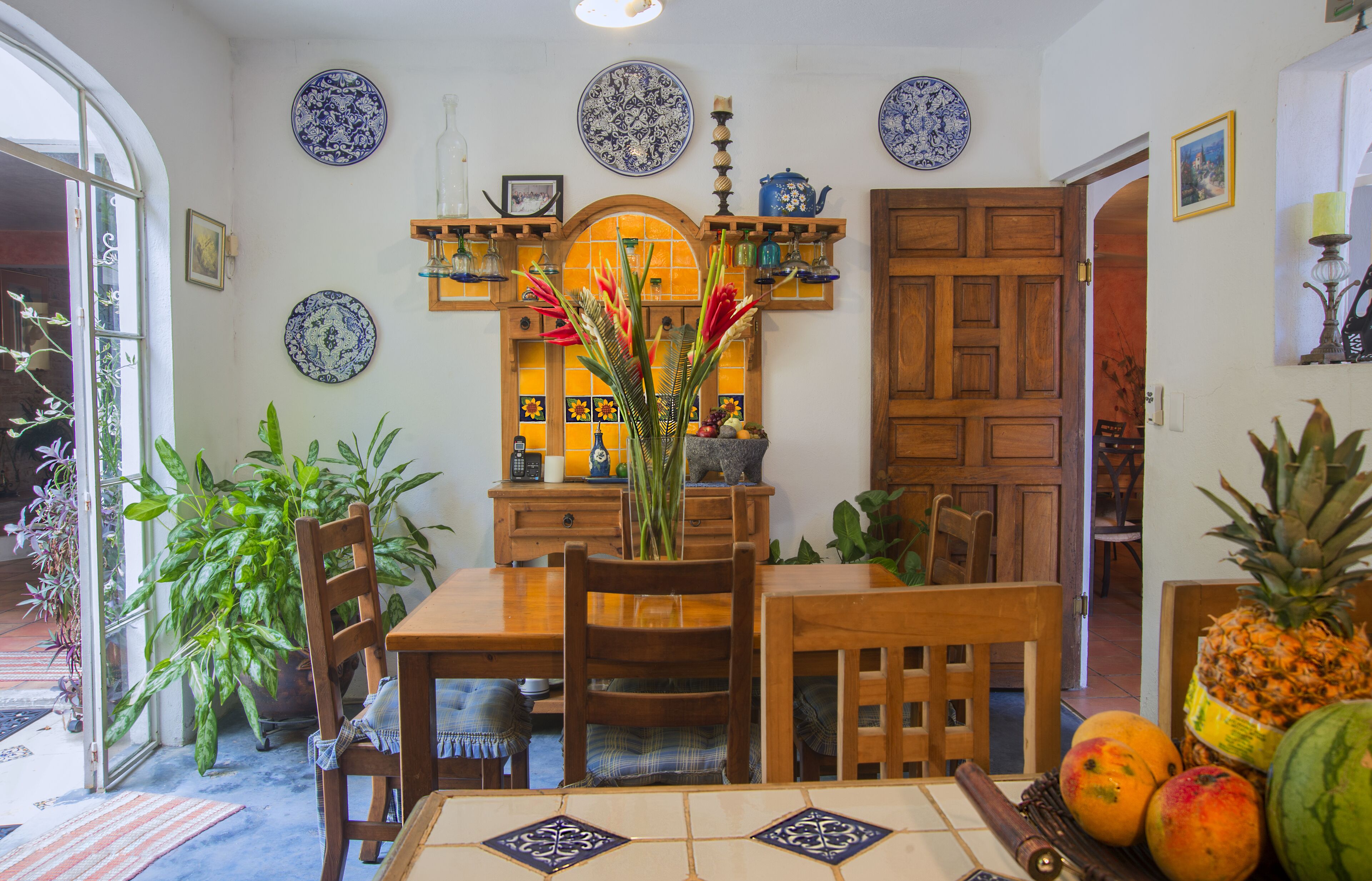 Un coin repas avec une table en bois, des chaises et un vase de fleurs. On peut également voir des plantes en pot et un panier de fruits sur la table.