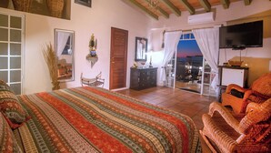 A spacious bedroom with a large bed, a sitting area with wicker furniture, a television, and a balcony with a view. - Ocean View - In the Heart of Old Town -Casa Romance Escondido (Puerto Vallarta)