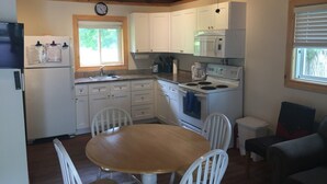 A compact kitchen with white cabinets, a microwave, stove, and a sink. There is a wooden dining table with chairs, and a clock on the wall. - Pine Cottage at Shiloh Cottages - Fenelon Falls (Fenelon Falls)