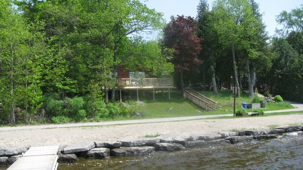 A dock extending into a body of water, surrounded by lush greenery and a wooden structure with a deck. - Pine Cottage at Shiloh Cottages - Fenelon Falls (Fenelon Falls)