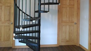 A spiral staircase with black railings and wooden steps, leading to a loft area with a wooden railing. - Pine Cottage at Shiloh Cottages - Fenelon Falls (Fenelon Falls)