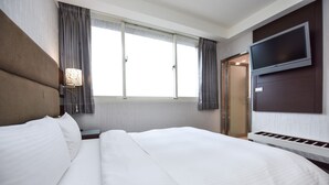 Habitación de hotel con una cama grande, un televisor de pantalla plana montado en la pared, una mesita de noche con lámpara y una ventana con cortinas.