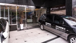Entrada de hotel con puerta de vidrio, una planta en maceta y un coche negro estacionado afuera.