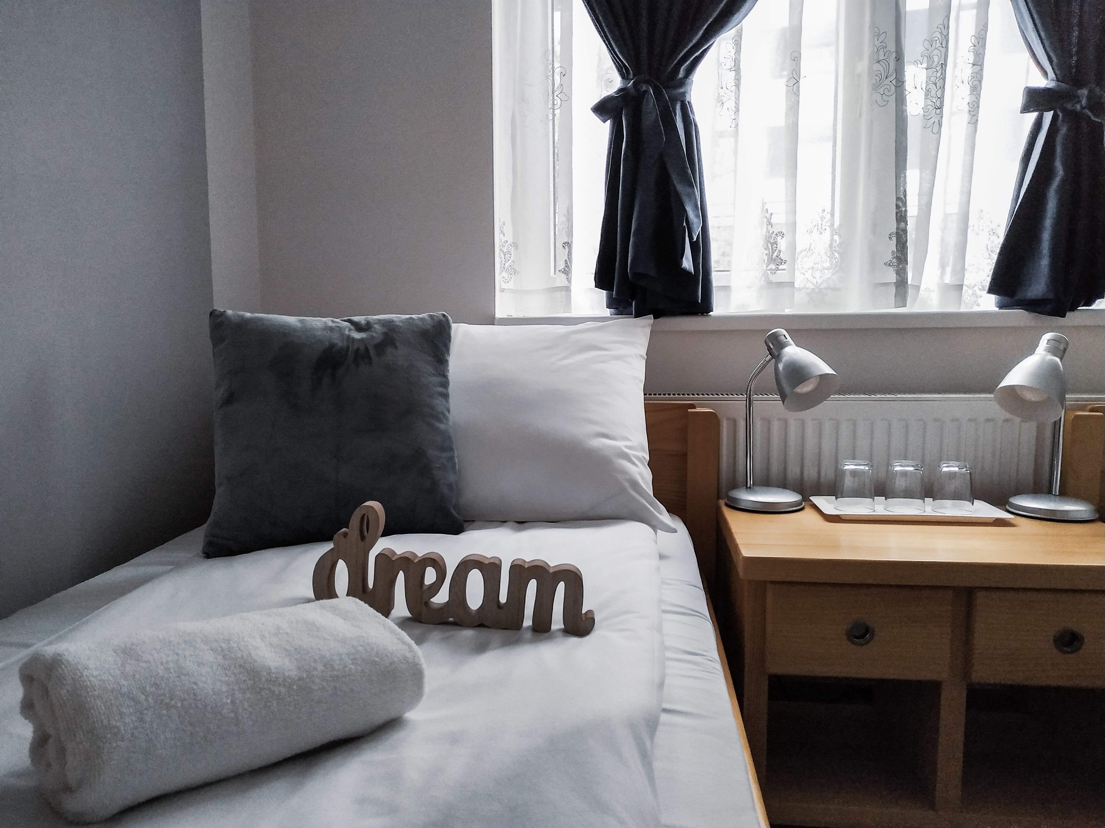 En snyggt bäddad säng med en kudde, en handduk och en träskylt med texten ”dröm”. Bredvid sängen står ett litet bord med en lampa och ett par glasögon.