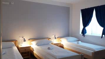 Een kamer met drie eenpersoonsbedden, elk voorzien van nachtkastje en lamp, een raam met gordijnen, en een stopcontact.