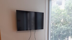 A small room with a wooden desk, a chair, a flat-screen TV mounted on the wall, a small white refrigerator, and a window with blinds. -  Bloom Inn (Niš)