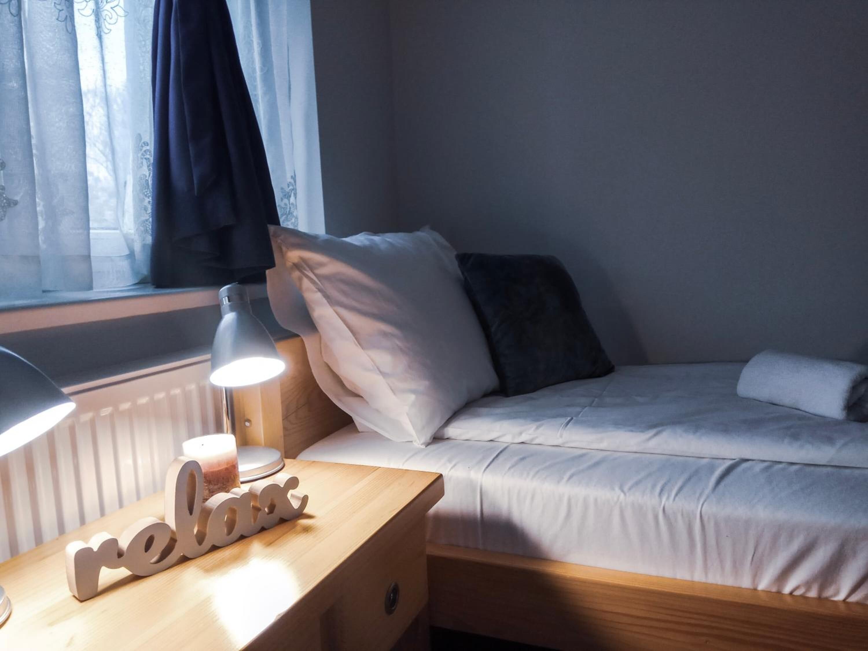 A neatly made bed with white bedding, a bedside lamp, a small wooden nightstand with a candle and a 'relax' sign, and a window with sheer curtains.