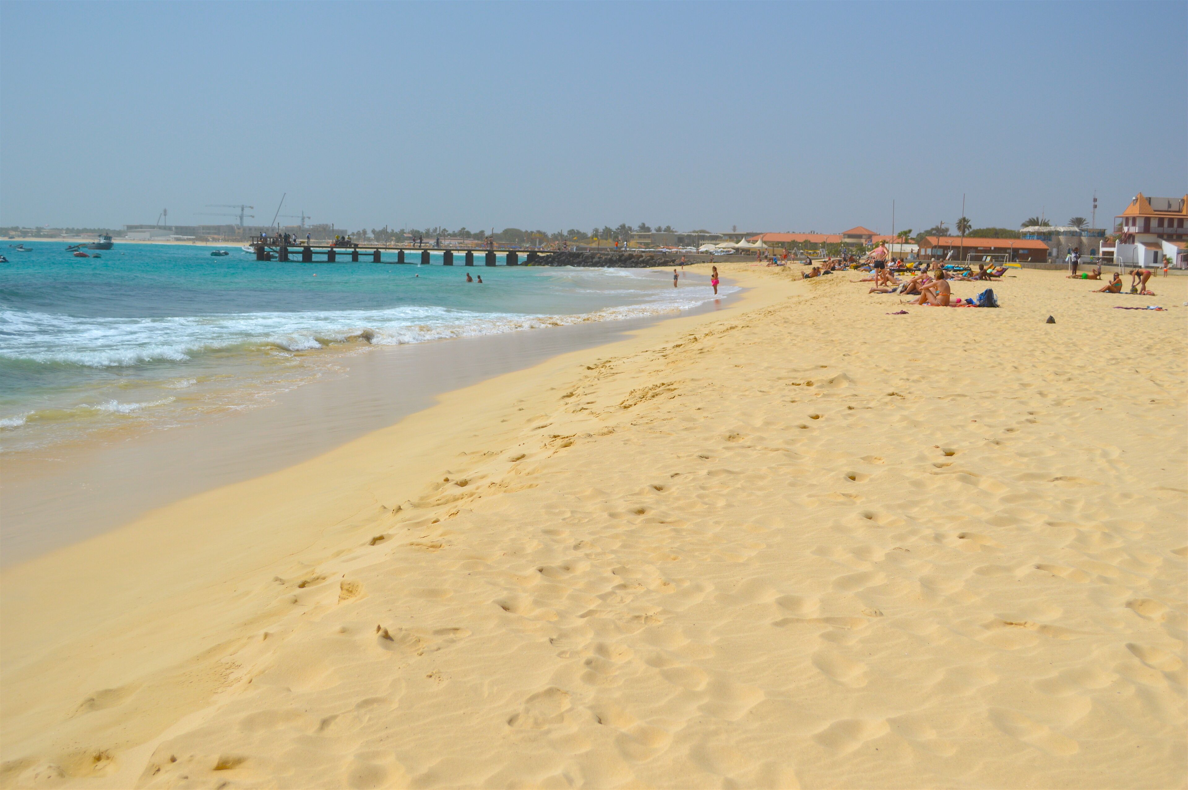 Une plage de sable avec des empreintes, une jetée et des bâtiments en arrière-plan.