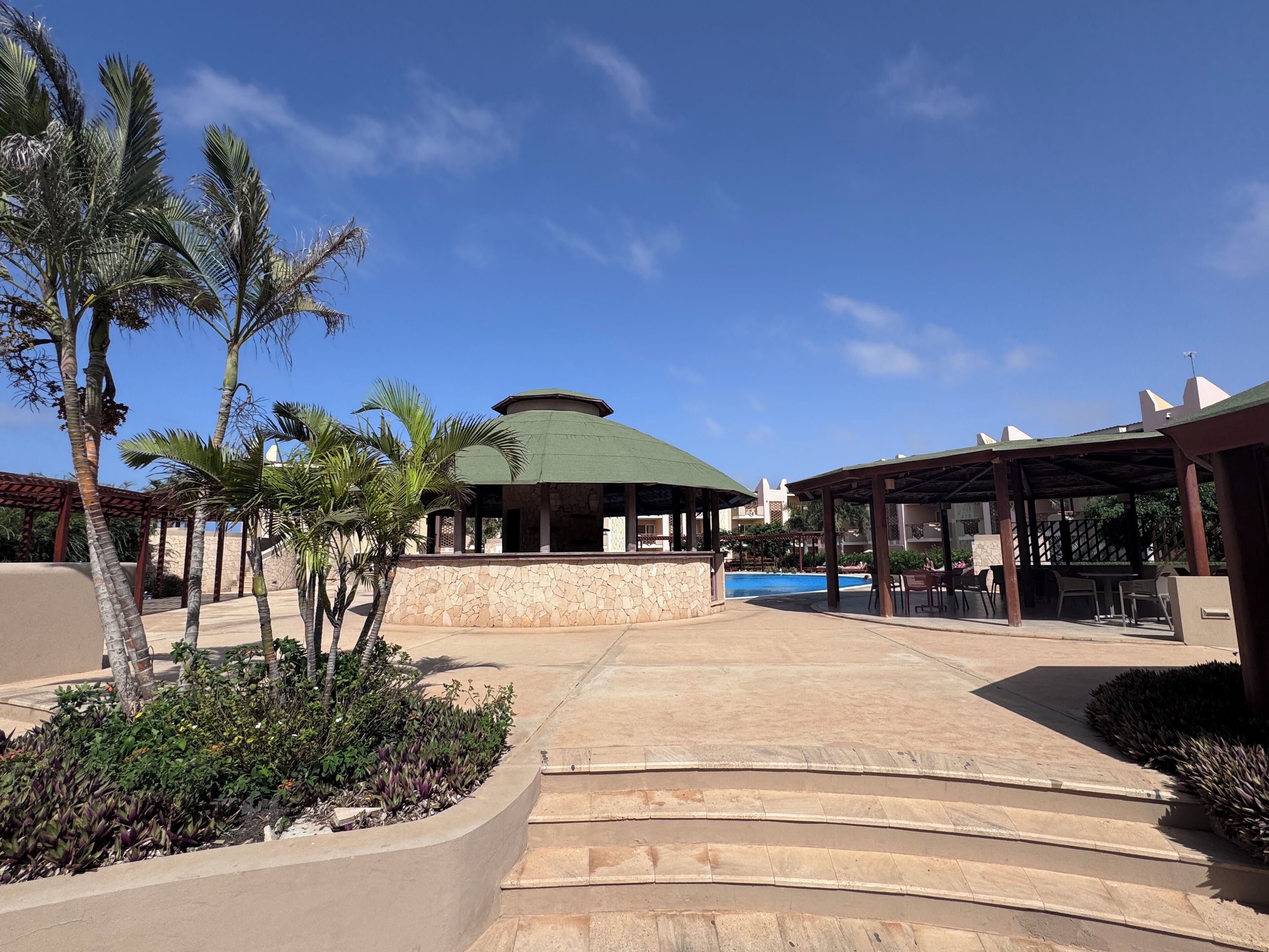 Zona de resort con piscina, zonas de estar cubiertas y palmeras.