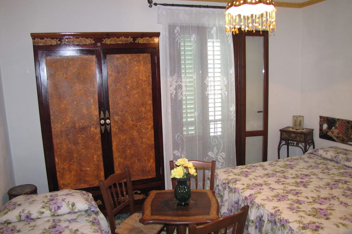 Ein Zimmer mit zwei Betten, einem Holztisch mit Blumen in einer Vase, einem Stuhl, einem Holzschrank, einem Spiegel und einer Kristallleuchter.