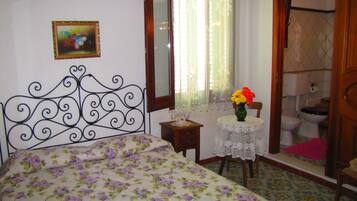 Una camera da letto con una coperta a fiori, un comodino con un vaso di fiori, uno specchio e una finestra con tende in pizzo.