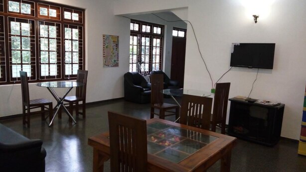 A room with a dining table, chairs, a sofa, a TV, and large windows with a view. - Enrich Bungalow (Kandy)