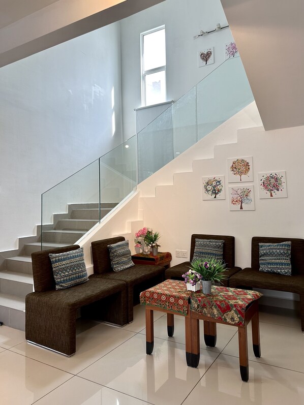 A modern living room with a glass staircase, two sofas with patterned cushions, a coffee table with a floral table runner, and wall art depicting trees.