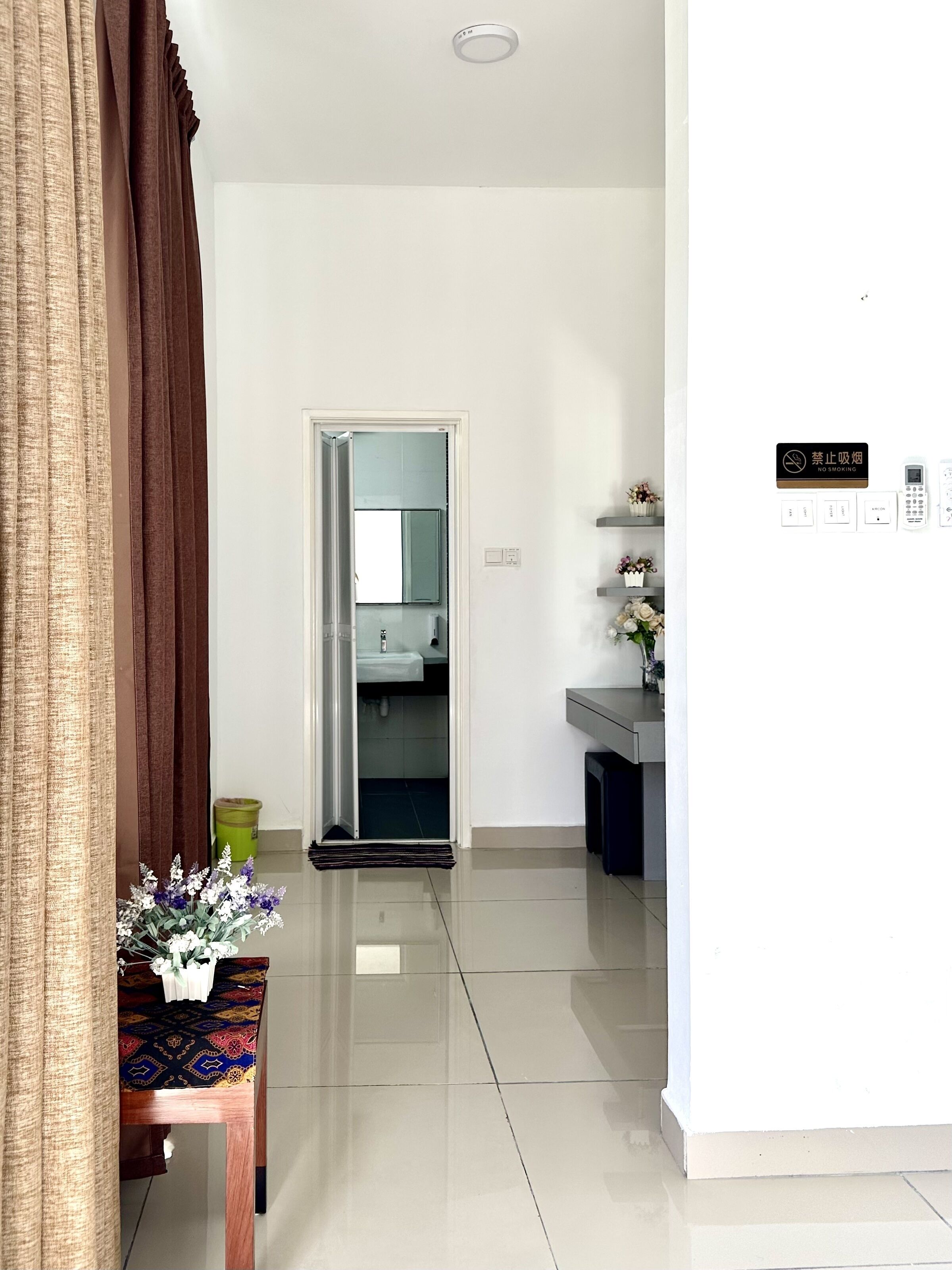 Une chambre d’hôtel moderne au design épuré et minimaliste. Elle est dotée d’un grand miroir, d’une petite table sur laquelle est posé un vase de fleurs, et d’un panneau « DÉFENSE DE FUMER » accroché au mur.