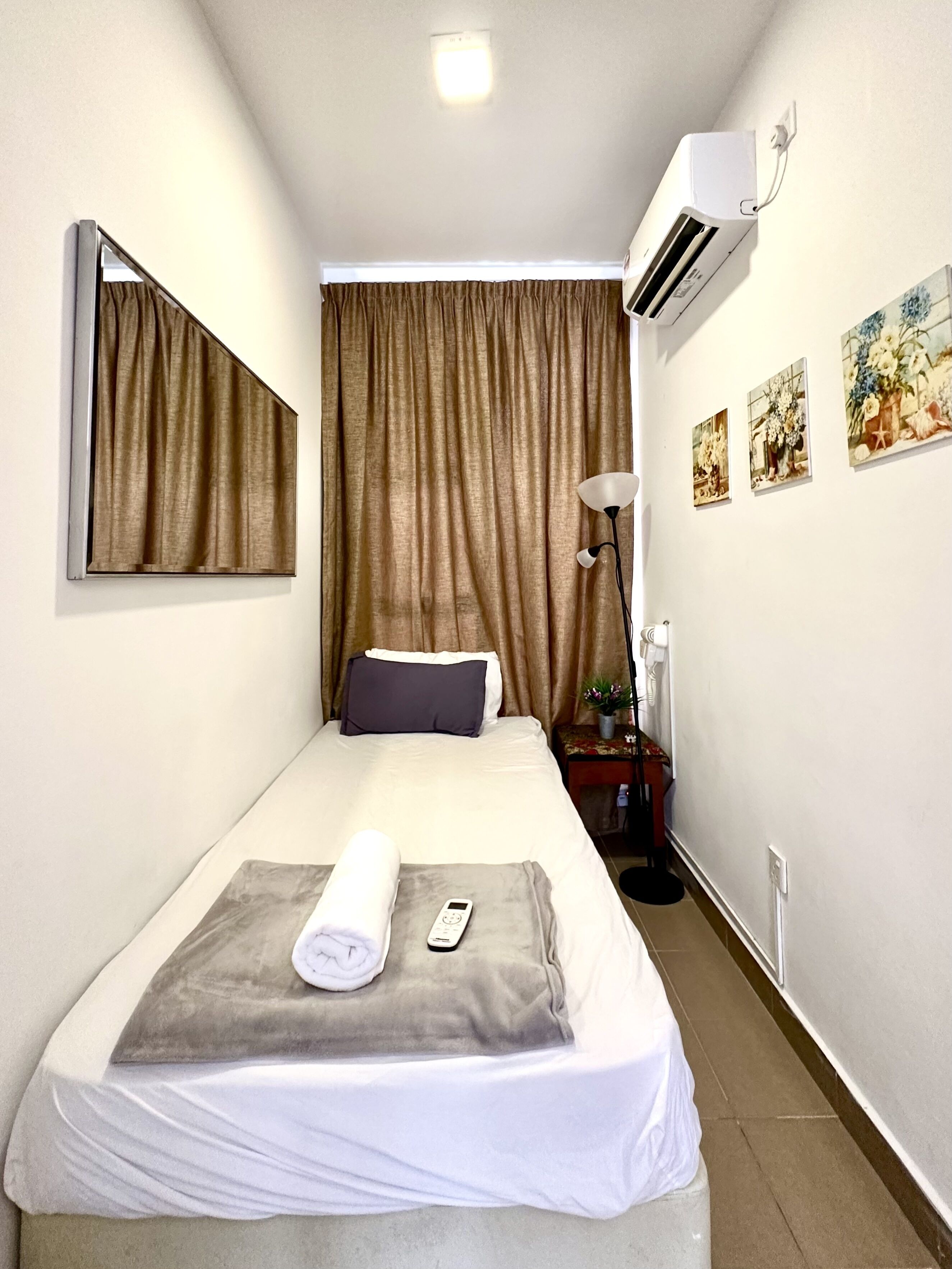 A hotel room with a bed, a bedside table with a lamp, a wall-mounted air conditioner, and framed artwork on the wall.
