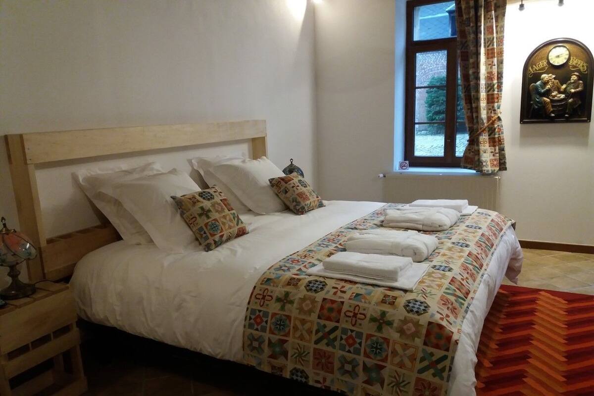 Uma cama bem arrumada com colcha estampada e almofadas, uma mesinha de cabeceira de madeira com abajur, e uma janela com cortinas.