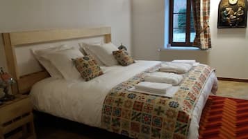 Un letto rifatto con una coperta a motivi e cuscini, un comodino in legno con una lampada, e una finestra con tende.