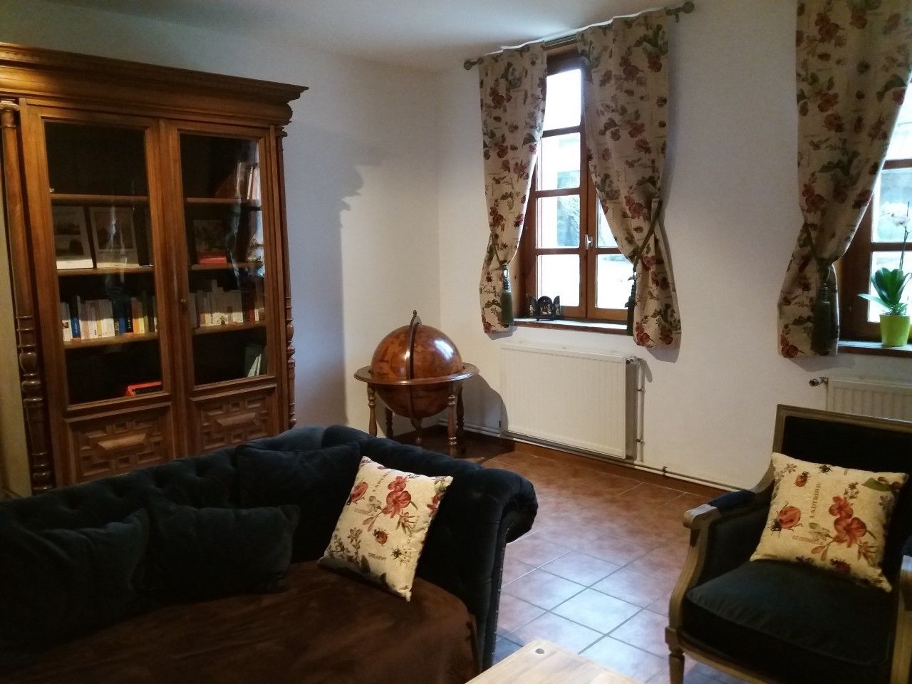 A living room with a dark sofa, armchair, wooden cabinet, globe on a stand, and floral curtains.