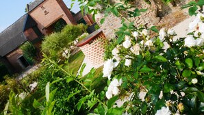 Um edifício de tijolos tradicional com telhado inclinado, rodeado por vegetação exuberante e plantas floridas brancas.
