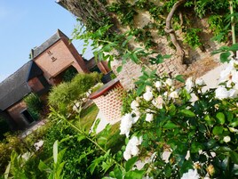 Un edificio de ladrillo tradicional con techo inclinado, rodeado de abundante vegetación y plantas con flores blancas.