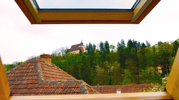 A view from a window with a skylight, overlooking a building with a red-tiled roof and a forested hill in the background. - Venesis House (Sighisoara)